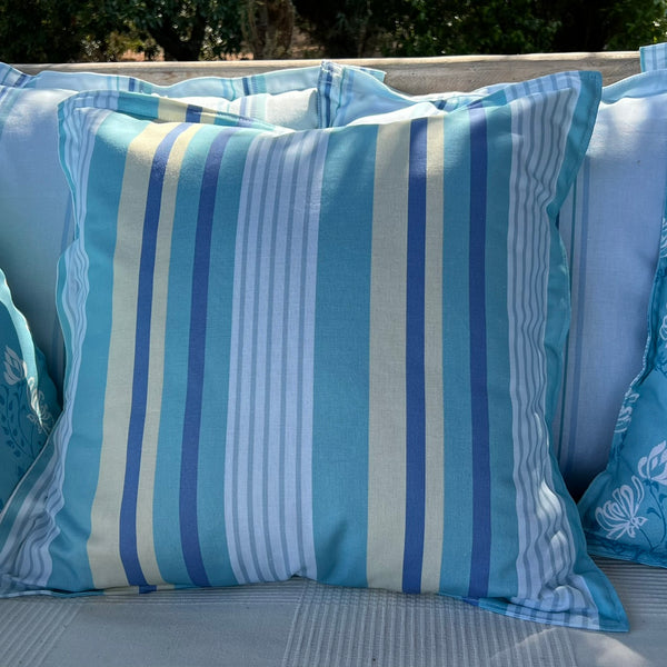 A square throw pillow with vertical stripes in light blue, medium blue, navy, and cream, displayed on a light-colored outdoor sofa with other pillows in the background. You can create your own colourway of this fabric.