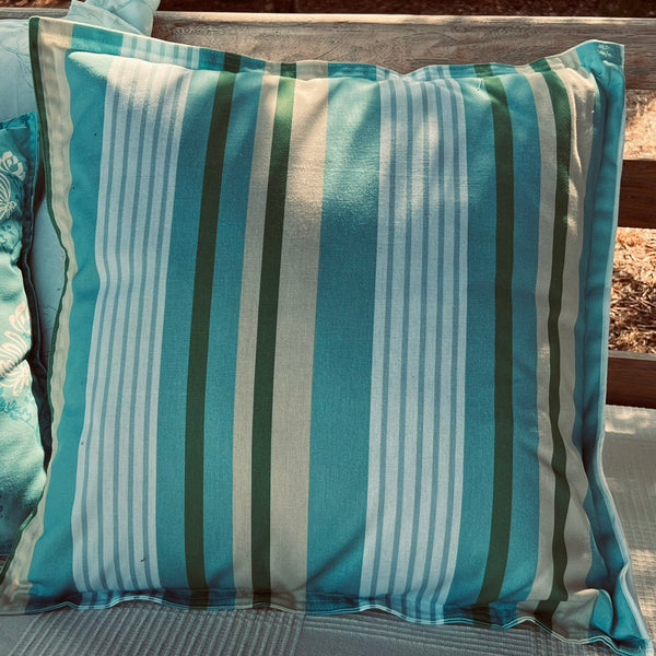 A square throw pillow which has been covered in a bespoke coloured vertical stripe cotton fabric. The colours used are teal, light blue, white, and dark green, resting on a wooden outdoor bench.