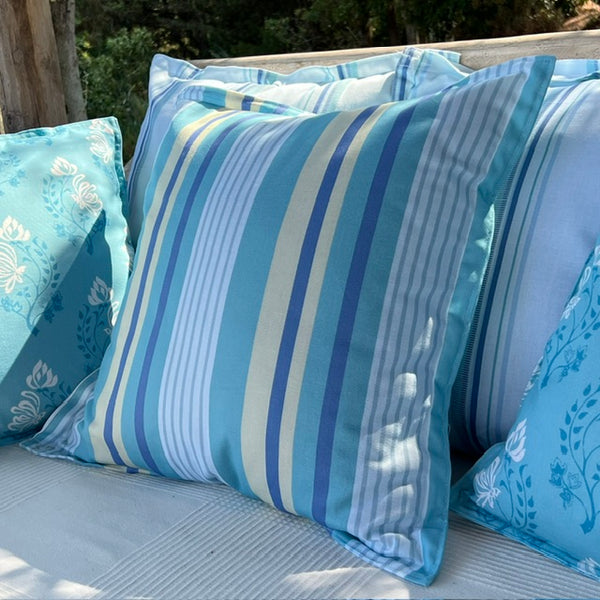 A set of custom coloured blue and white striped outdoor throw pillows displayed on a cushioned bench with a natural wooden post and green foliage in the background.