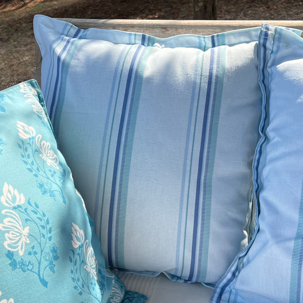 A light blue striped outdoor throw pillow custom printed in tones of blue shown alongside other blue patterned pillows on a patio bench.